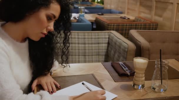Χαριτωμένο μελαχρινή κοπέλα με τα σγουρά μαλλιά γράφοντας ένα ποίημα σε ένα εστιατόριο — Αρχείο Βίντεο