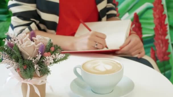 La mano di una giovane donna in abito a righe prende appunti con una matita in un blocco note rosso — Video Stock