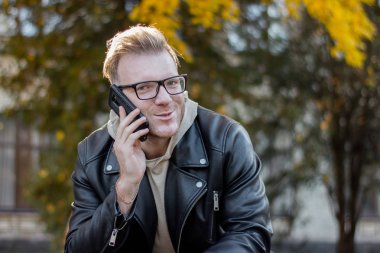 Rahat giysiler Içinde Avrupa gülümseyen akıllı adam telefonda konuşuyor ve bir bankta oturuyor