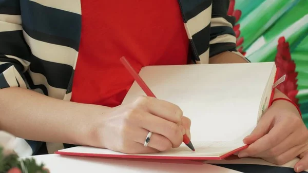 A mão de uma jovem mulher em um terno listrado toma notas com um lápis em um bloco de notas vermelho — Fotografia de Stock