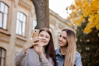 Üniversite yakınlarındaki sonbahar parkında telefonda selfie çeken iki güzel genç kız