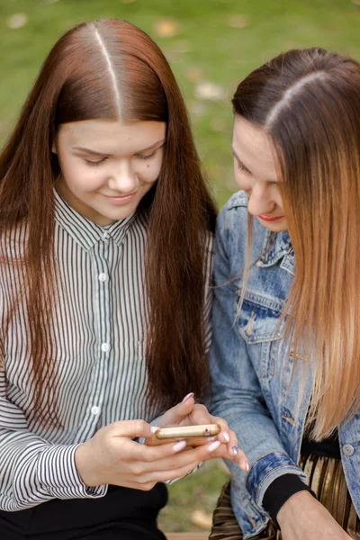 Retrato de duas meninas bonitas estão olhando para o telefone, sentado em um banco no parque de outono — Fotografia de Stock