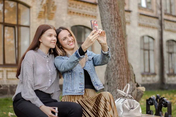 Zwei schöne junge Mädchen machen ein Selfie am Telefon, während sie auf einer Bank im herbstlichen Park in der Nähe der Universität sitzen — Stockfoto