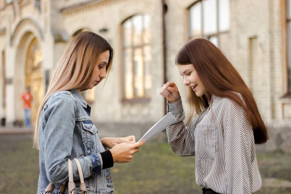 Duas namoradas ficam perto da universidade e olham para os jornais. — Fotografia de Stock