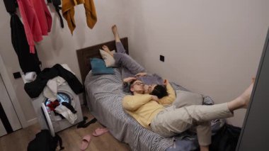 Bir çift yumuşak bir yatakta dinleniyor. Ayakları havada, dağınık yatak odasında rahat bir atmosfer yaratıyorlar, rahatlamayla dolu. Çevreleri neşeli ev atmosferini arttırır.