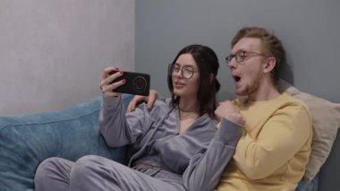 Gözlüklü mutlu çift kanepeye oturur ve akıllı telefonuyla bir selfie çeker. Neşeli atmosfer onların neşeli bağlantılarını sembolize eder. Mutluluklarını ve sevgilerini gösterir.