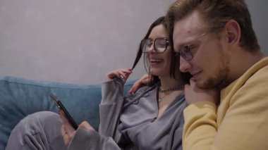 Sıcak ve samimi bir çift gözlük takıyor koltukta yan yana oturuyor, telefonda komik videolar seyrediyor, mutluluk ve sıcaklık saçıyor, misafirperver, rahatlatıcı bir atmosferde