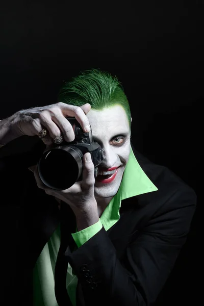 Portrait of a joker. Makeup for Halloween. Crazy image of a photographer man in a green shirt with green hair with camera in hand