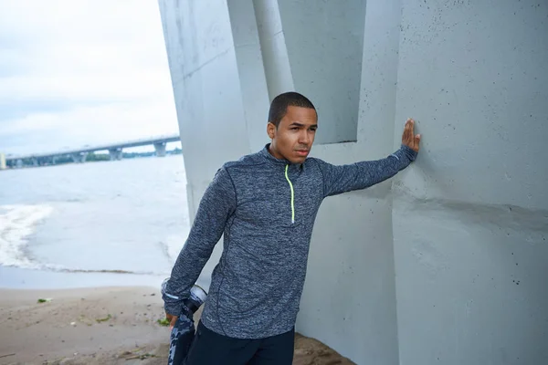 Picture of black young male jogger dressed in trendy outfit stretching legs before running exercise outdoors, leaning on grey wall of concrete construction, having determined confident look