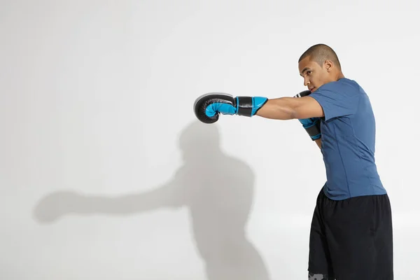 Profile studio shot of determined young dark skinned man boxer with shaven head working out in gym, practicing punches, posing at white wall, stretching out his left hand. Martial arts concept