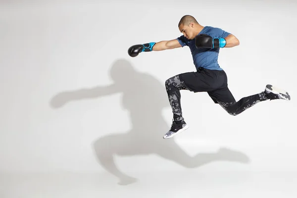 Full length sideways portrait of fit atheltic young African American male kickboxer wearing stylish outfit and boxing gloves jumping and throwing punch, casting shadow upon white studio wall