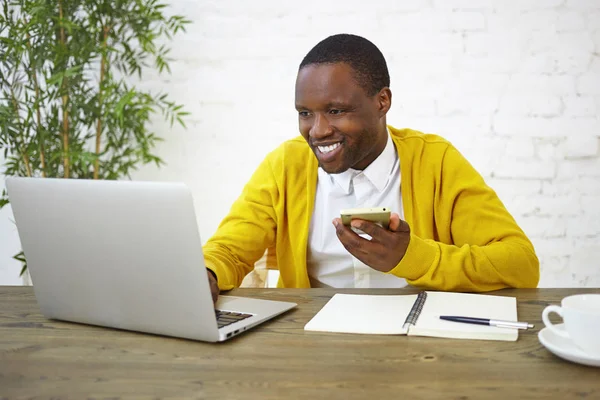 People, lifestyle, job, technology and communication concept. Cheerful Afro American male freelancer in bright yellow cardigan using cell phone and portable computer in home office, smiling broadly