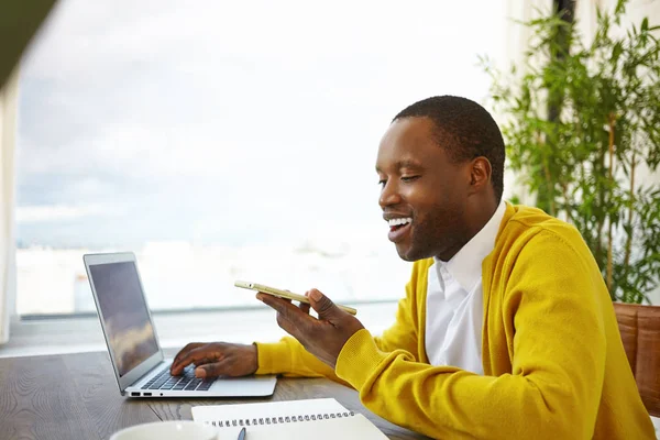 Sideways shot of stylish young dark skinned male enjoying high speed wifi internet on his modern electronic gadgets, using laptop at cafe during coffee break, recording audio file on smart phone