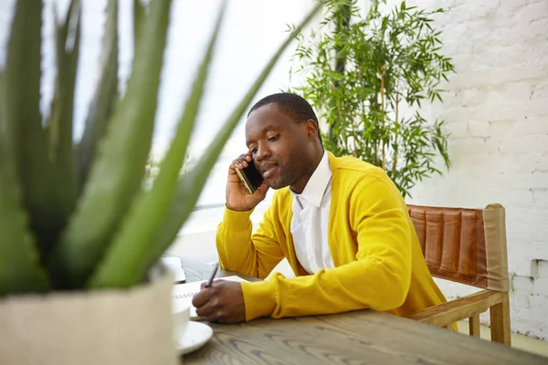 Handsome successful young dark skinned businessman making notes in his copybook while speaking on mobile phone to business partner, sitting at office, surrounded with plants. Selective focus