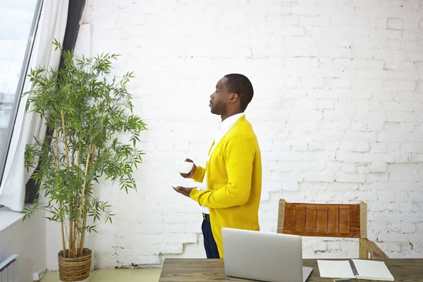 Stylish young African male creative worker or businessman in yellow cardigan drinking fresh morning coffee, enjoying view from office window, standing at his workplace with laptop and copybook on desk