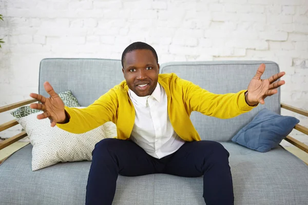 Indoor shot of attractive adult dark skinned man sitting on couch in living room and gesturing emotionally, keeping hands wide while watching football game on TV, supporting his favorite team
