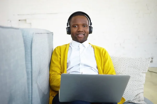 Handsome black male student wearing yellow cardigan over white shirt studying at home, using laptop and headphones, listening to lecture online. Happy man enjoying music via headset on sofa