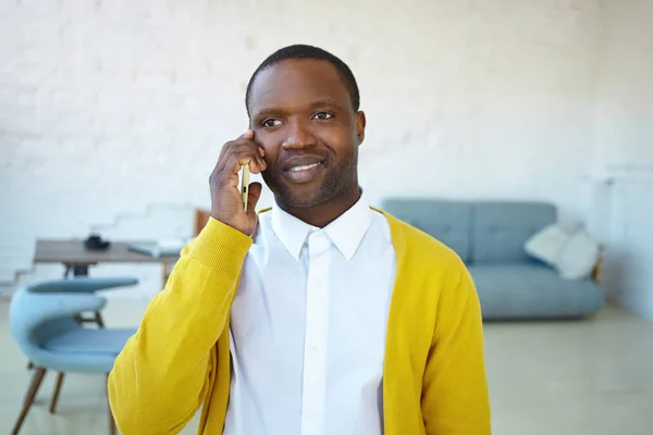 Horizontal shot of handsome cheerful African young man wearing white shirt and yellow cardigan, smiling broadly while having nice phone conversation, talking to friend, receiving good positive news