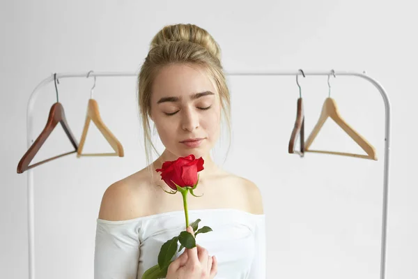 Beautiful young female with hair bun and naked shoulders posing in studio with empty hangers in background, keeping eyes closed, enjoying sweet fresh aroma coming form red rose in her hands