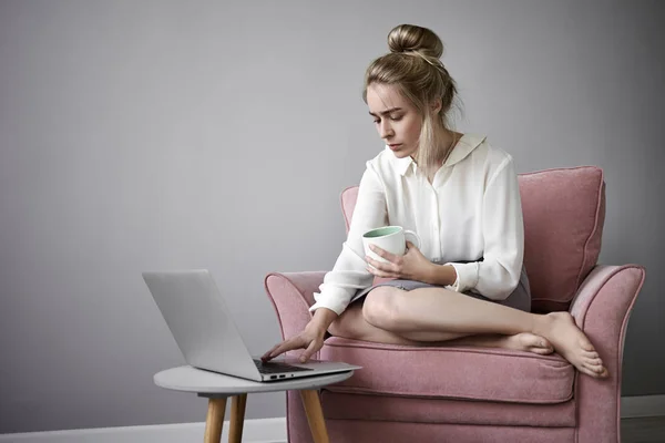Attractive female freelancer in white blouse working remotely on portable computer, sitting barefooted in armchair, drinking hot tea or coffee, keeping one hand on keyboard, having focused look