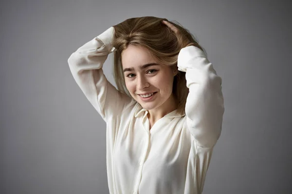 Horizontal shot of positive cheerful young woman wearing elegant white blouse smiling joyfully at camera, touching her long hair, rejoicing at new hairstyle. People, fashion and style concept