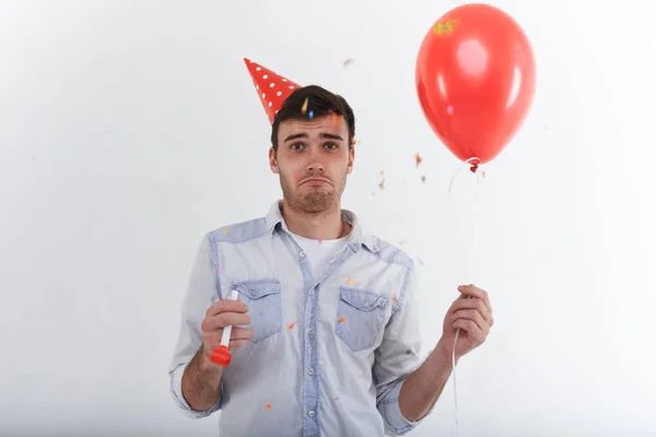 Human emotions. Sad unhappy young European male with stubble having displeased disappointed look, feeling lonely on his birthday party, holding red party horn and helium balloon in his hands