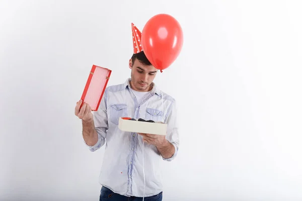 Picture of curious young man with stubble posing in studio with helium balloon and opened present box. Attractive birthday guy on party opening box, having anticipating look People and celebration
