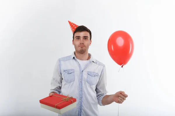 Horizontal shot of funny young Caucasian man in denim shirt holding red balloon and birthday present in fancy box, looking at camera with disappointed expression on his face. Human emotions