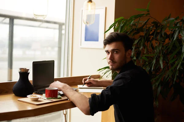 Modern dressed man sit in cafe with notebook and look to the camera with hands on laptop. Freelancer wait for the client for new job and use free wi-fi to surfing internet pages.