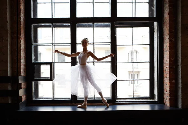 Full lenght picture of female classical ballet dancer in white tutu dancing in ballet studio, standing on wide windowsill and looking at camera. People, profession, hobby and occupation concept