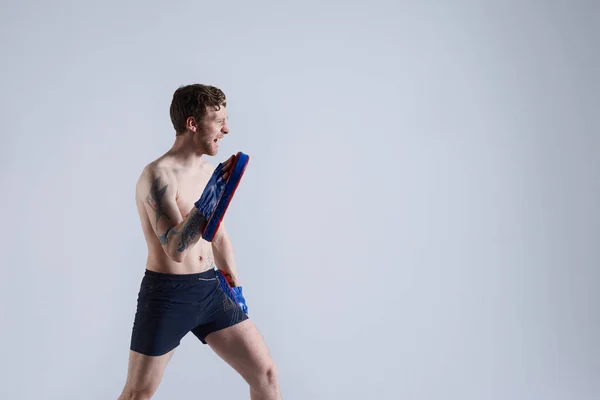 Profile portrait of adult bearded professional boxer with muscular body training boys, giving them instructions, wearing shorts and boxing punch mitts, standing against white wall background