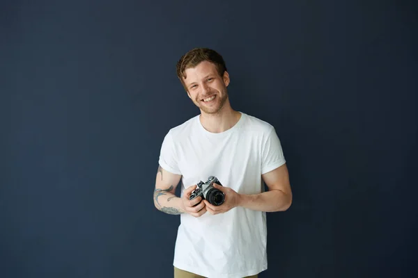 People, hobby, leisure, job and occupation concept. Cheerful handsome guy with tattooed arm smiling broadly, holding photo camera in his hands, taking pictures of people and events around him