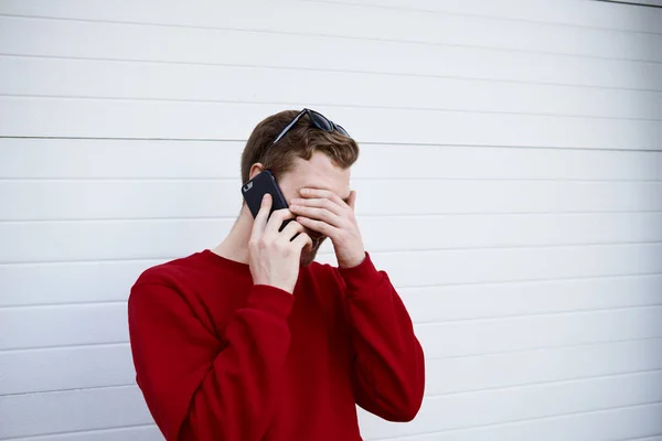 Bearded young Caucasian male in sweater having phone conversation with his friend, making facepalm gesture while laughing at funny story or joke. Modern technology and communication concept