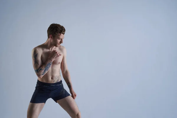 Profile picture of handsome sportsman with beard and tattooed arm wearing trunks standing shirtless in defensive posture while practicing in gym, working out skills and moves. Martial arts concept