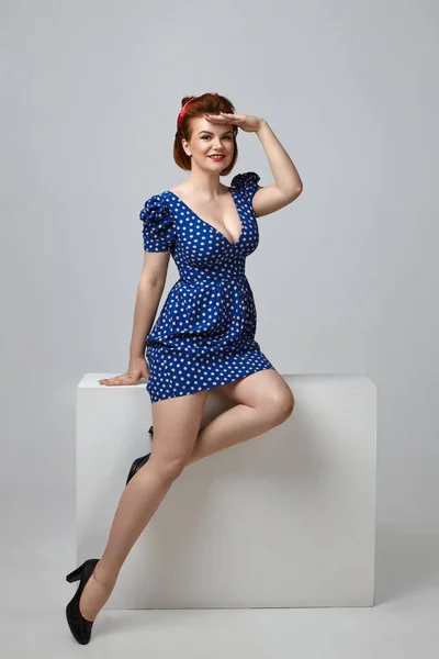 Glamourous young lady with big breast and ginger hair sitting comfortably in studio, holding hand at forehead, trying to see something.