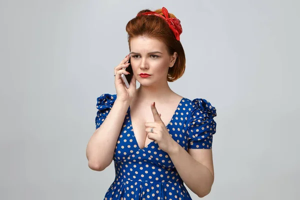 Angry young woman in vintage outfit having strict look, raising index finger in front of, talking on smartphone.