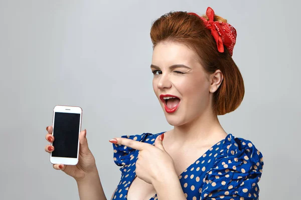 Emotional young woman dressed in retro style winking playfully at camera and pointing at smartphone.