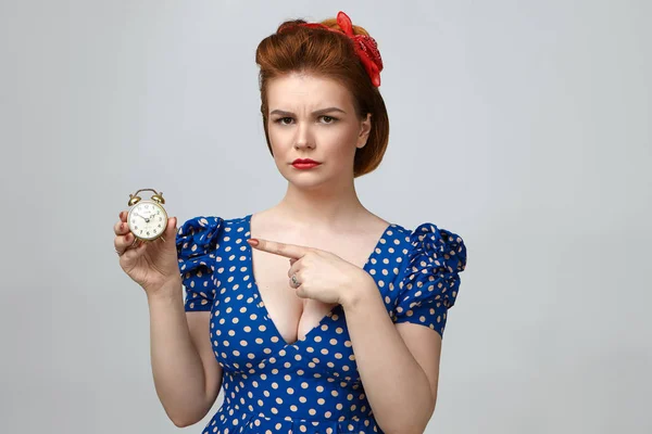 Studio shot of angry young woman dressed in vintage clothes holding alarm clock, looking at camera with annoyed expression, pointing index finger.