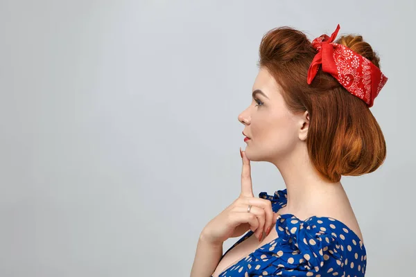 Pensive young woman in dotted blue dress and red headband keeping finger on chin.