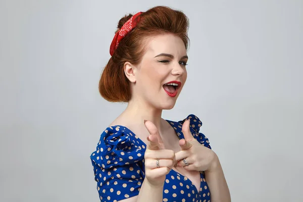 Attractive woman wearing retro hairstyle, make up and vintage dress winking and pointing two index fingers at camera, flirting.