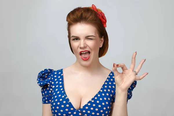 Emotional attractive young woman wearing retro outfit and bright makeup having playful flirty look, winking and making ok sign at camera.