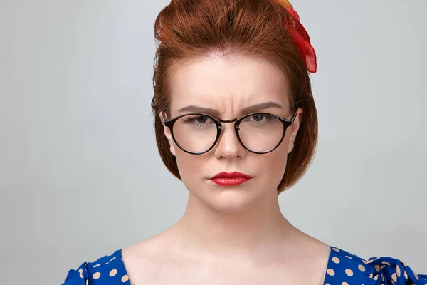 Attractive young female teacher wearing dotted dress, red lipstick and stylish eyeglasses frowning, looking at camera with strict expression.