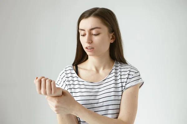 Young woman in striped top frowning, holding hand on aching wrist, massaging pain area, having painful facial expression.