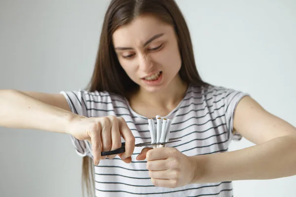 Angry young woman in striped t-shirt cutting bunch of cigarettes in halves using scissors, having fierce facial expression.