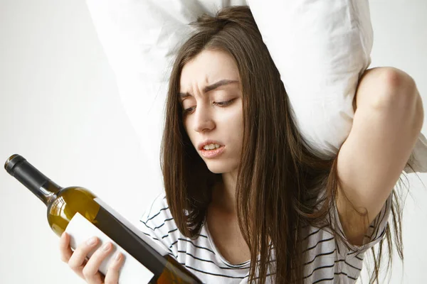 Stressed frowning young woman in striped pajamas holding pillow on head and looking at empty bottle, feeling desperate, suffering from hangover after party.
