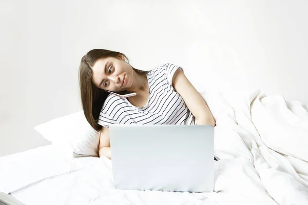 Serious young woman using generic notebook pc and smartphone lying in bed, having concentrated focused facial expression.