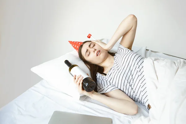 Party hard concept. Frustrated stressed young woman lying in white bed with bottle of wine, touching face, having headache and suffering from hangover after birthday party last night.