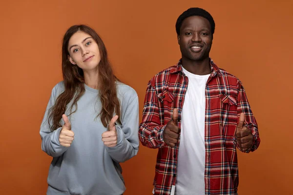 Positive cheerful young afro american guy and cute long haired caucasian woman enjoying time together, smiling joyfully and showing thumbs up gestures