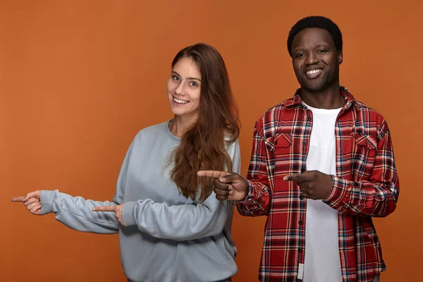 Picture of emotional overjoyed young afro american guy posing in studio with adorable friendly looking caucasian girlfriend, pointing fore fingers sideways and smiling broadly.