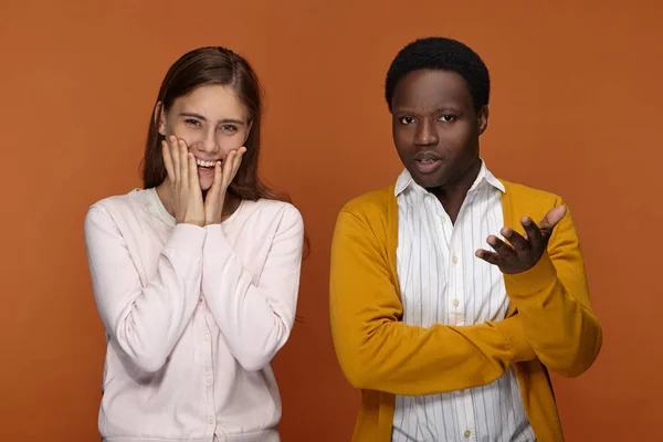 Confused clueless young african american man gesturing, being at loss, positive caucasian woman holding hands on cheeks and smiling happily, feeling excited.